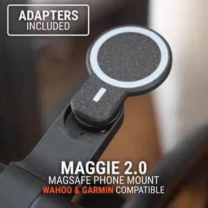 Op de afbeelding: Een zwarte MagSafe telefoonhouder met een cirkelvormig magnetisch kussen en een witte rand. De houder is voorzien van het label "MAGGIE 2.0" en "WAHOO & GARMIN COMPATIBLE." De tekst "ADAPTERS INCLUDED" is ook zichtbaar.