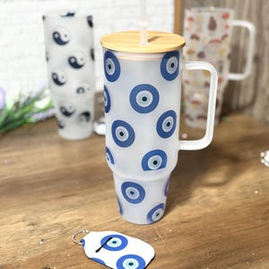 May include: A frosted glass tumbler with a handle, a bamboo lid, and a clear straw, featuring a repeating blue and white evil eye pattern. In the foreground, a matching white keychain with a blue evil eye design and a metal ring is visible.