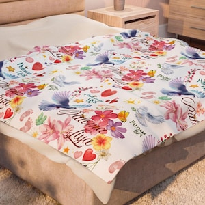 May include: A white blanket featuring a repeating pattern of colorful flowers, blue birds, red hearts, and small footprints. The text Paloma Liya is printed in a cursive script throughout the design, interspersed with yellow stars and green leaves.