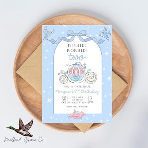 May include: A light blue birthday invitation with the text "Bibbidi Bobbidi Two" and a drawing of a carriage. The invitation is for Morgan's 2nd birthday, with the date, time, and location details. The invitation is on a wooden tray with an envelope.