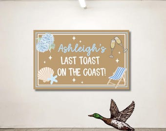 May include: A rectangular sign with the text "Ashleigh's Last Toast on the Coast!" in white and light blue. The sign features illustrations of a hydrangea, seashells, champagne glasses, and a beach chair. A flying duck is in the foreground.