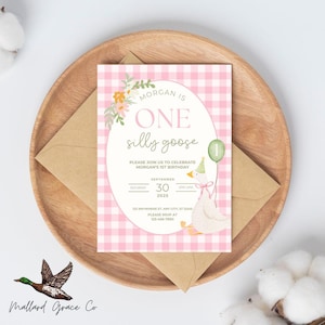 May include: A birthday invitation with a pink gingham background and the words "MORGAN IS ONE silly goose." The invitation features a watercolor illustration of a goose holding a balloon. The invitation is on a wooden plate with a brown envelope.