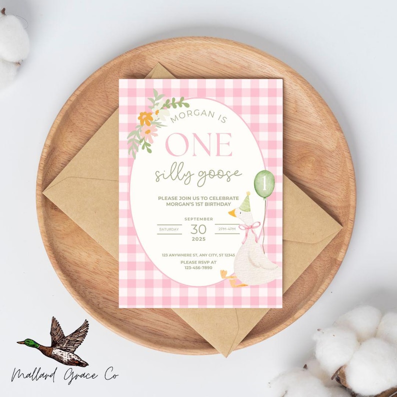 May include: A birthday invitation with a pink gingham background and the words "MORGAN IS ONE silly goose." The invitation features a watercolor illustration of a goose holding a balloon. The invitation is on a wooden plate with a brown envelope.