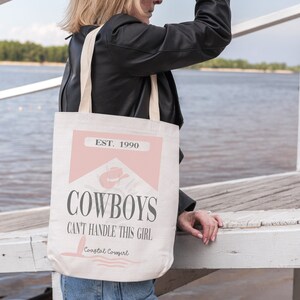 K&ouml;nnte beinhalten: Eine wei&szlig;e Canvas-Tragetasche mit einem rosa-wei&szlig;en Design. Die Tasche tr&auml;gt den Text "EST. 1990" und "COWBOYS CAN'T HANDLE THIS GIRL" mit einem rosa Cowboyhut-Motiv. Der Text "Coastal Cowgirl" ist in kleinerer Schrift am unteren Rand der Tasche aufgedruckt.