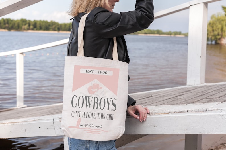 K&ouml;nnte beinhalten: Eine wei&szlig;e Canvas-Tragetasche mit einem rosa-wei&szlig;en Design. Die Tasche tr&auml;gt den Text "EST. 1990" und "COWBOYS CAN'T HANDLE THIS GIRL" mit einem rosa Cowboyhut-Motiv. Der Text "Coastal Cowgirl" ist in kleinerer Schrift am unteren Rand der Tasche aufgedruckt.