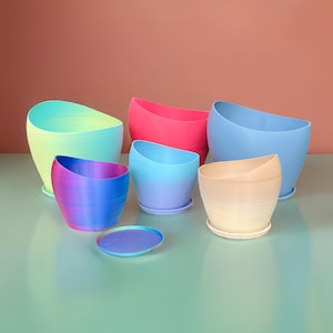 May include: A collection of colorful, modern planters in various sizes and shades, including blue, pink, and beige. Each planter has a unique, angled design and sits on a matching saucer. The planters are arranged on a light green surface against a peach background.
