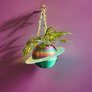 Könnte beinhalten: Ein hängender Pflanzentopf in Planetenform mit einem regenbogenfarbenen Körper und einem weißen Ring. Er enthält eine grüne Pflanze und wird an einem Seil vor einer violetten Wand aufgehängt.