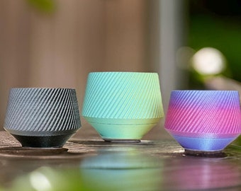 May include: Three small, decorative planters with a geometric design. One is black and gray, another is light green and blue, and the third is pink and purple. The planters are sitting on a wooden surface, with green foliage in the background.