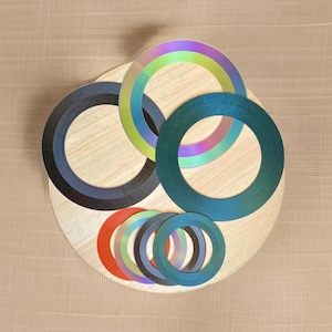 May include: A collection of colorful, iridescent, and solid-colored circular shapes arranged on a light wood-grain surface. The circles vary in size and color, including blue, green, red, and rainbow hues. The arrangement creates a layered, artistic composition.