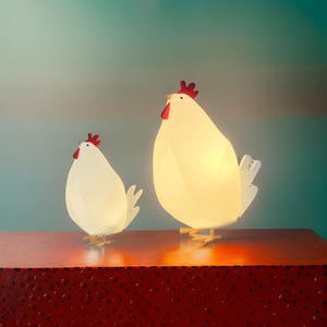 Peut inclure: Deux lampes en forme de poulet blanc avec des crêtes rouges et des pieds jaunes sont exposées sur une surface rouge. Les lampes sont éclairées, créant une lueur chaude. L'arrière-plan est un dégradé de bleu sarcelle et de bleu.