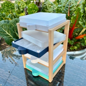 May include: A multi-tiered seed starting tray system. The wooden frame holds four clear plastic trays with colored handles in white, navy, mint green, and clear. The trays are designed for starting seeds indoors.