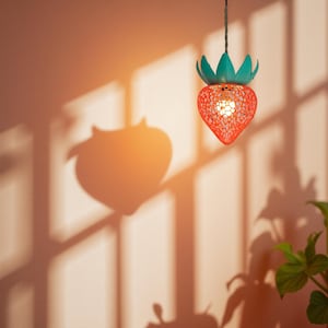 Op de afbeelding: Een hangende aardbeivormige lamp met een rood, opengewerkt ontwerp en een groene kroon. De lamp is van binnenuit verlicht en geeft een warme gloed. De schaduw van de lamp is zichtbaar op de perzikkleurige muur.