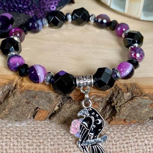 May include: A purple and black beaded bracelet with a silver charm of a raven holding a pink rose. The bracelet is made with agate and faceted black beads.
