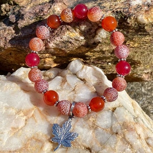 May include: A red agate gemstone bracelet with a silver maple leaf charm. The bracelet is made of 8mm beads and has a white and red agate pattern.