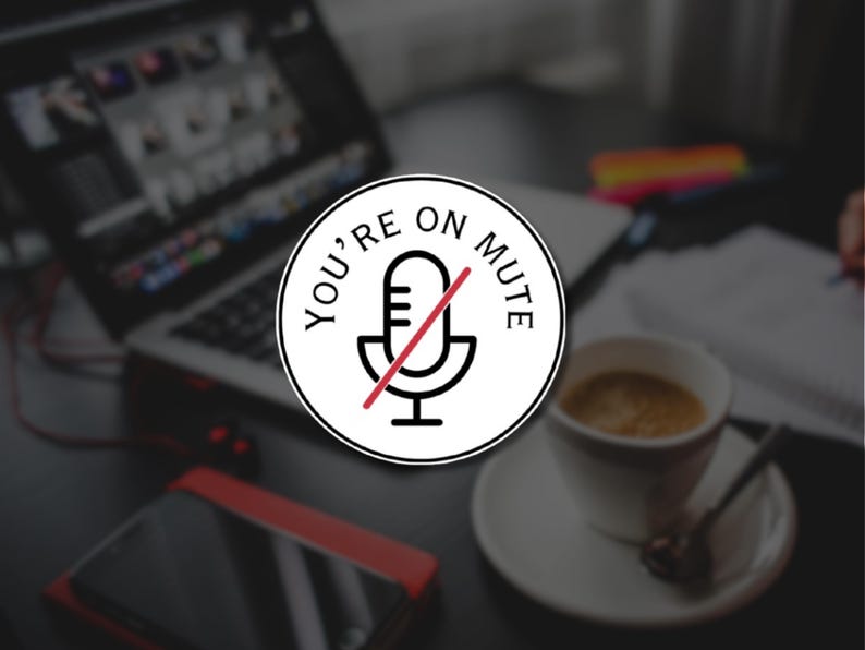 May include: A white circular sticker with the text "YOU'RE ON MUTE" and a microphone icon crossed out in red. The sticker is on a desk with a laptop, coffee cup, and smartphone. The image suggests a work-from-home or online meeting theme.