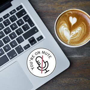 May include: A laptop with a keyboard and a round sticker that says "YOU'RE ON MUTE" with a microphone icon. A cup of coffee with latte art is next to the laptop on a wooden surface.