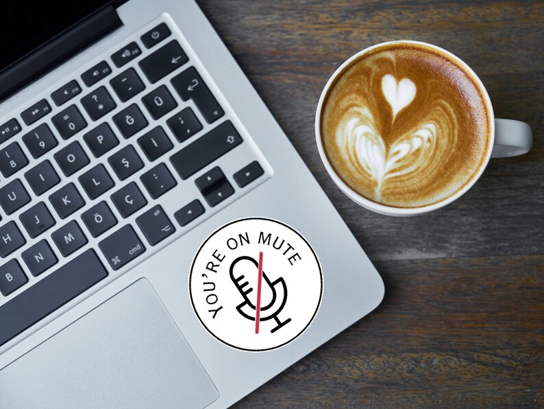 May include: A laptop with a keyboard and a round sticker that says "YOU'RE ON MUTE" with a microphone icon. A cup of coffee with latte art is next to the laptop on a wooden surface.