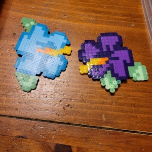 May include: Two pixel art flower designs made with colorful perler beads. One flower is blue with yellow accents and the other is purple with orange accents.