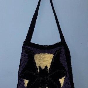 May include: A handmade crochet shoulder bag featuring a black bat design on a cream-colored background. The bag has a dark purple body, a black border, and a black strap. The bag is hanging against a light blue wall.