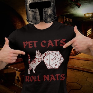 May include: A black t-shirt with the text "PET CATS ROLL NATS" in red. A white cat is depicted holding a red 20-sided die.