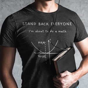May include: A man wearing black headphones and glasses is holding a black notebook. He is wearing a black t-shirt with the text "Stand Back Everyone I'm about to do a math problem" and a graph.