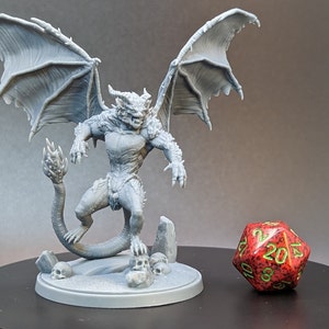May include: A gray 3D printed miniature of a winged demon with a long tail, standing on a base with skulls. The miniature is posed in a threatening stance with its wings spread wide. A red 20-sided die is in the foreground.