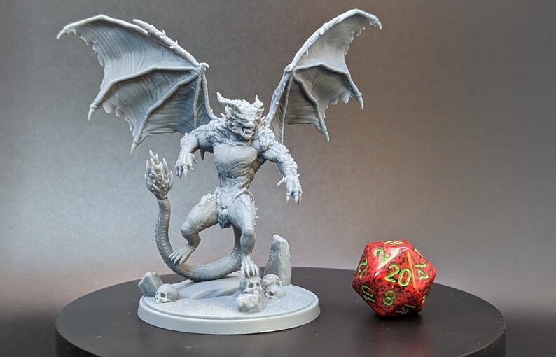 May include: A gray 3D printed miniature of a winged demon with a long tail, standing on a base with skulls. The miniature is posed in a threatening stance with its wings spread wide. A red 20-sided die is in the foreground.