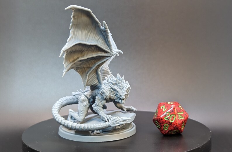 May include: A gray, 3D printed miniature dragon with wings spread, perched on a round base. The dragon has a spiky head and a long tail. A red 20-sided die is in the background.
