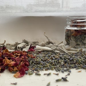 May include: A glass jar filled with dried herbs and flowers, including white sage, lavender, and rose petals. The jar is sitting on a white surface with loose herbs and flowers scattered around it.