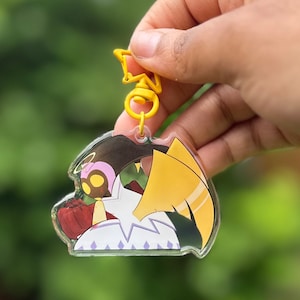 Könnte beinhalten: Ein durchsichtiger Acryl-Schlüsselanhänger mit einer Comicfigur mit gelben, grünen und roten Akzenten. Die Figur hat einen Heiligenschein und Flügel. Der Schlüsselanhänger hat einen gelben Metallverschluss.