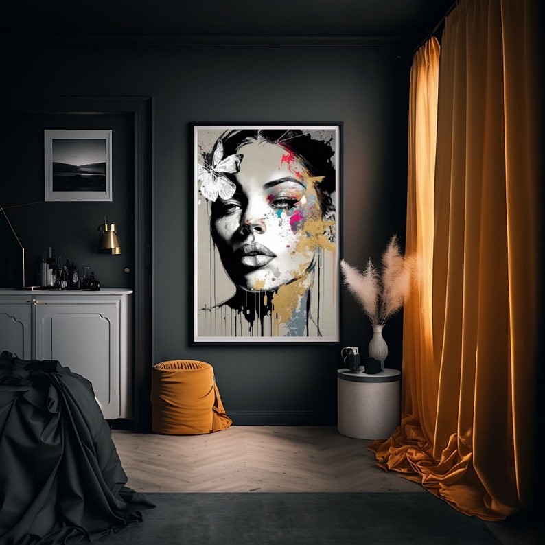 May include: A framed art print featuring a woman's face with abstract paint splatters in gold, pink, and blue. A white butterfly adorns her hair. The artwork hangs on a dark gray wall in a room with a yellow ottoman and curtains.