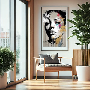 May include: A framed art print featuring a woman's face with a butterfly and colorful paint splatters. The artwork is in black, white, gold, pink, and blue. The print hangs above a wooden chair with a gray cushion, next to a large potted plant.