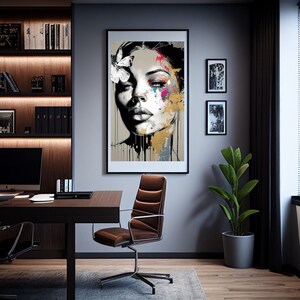 May include: A framed art print featuring a woman's face with a butterfly and colorful paint splatters. The artwork is in a black frame and hangs on a gray wall. The room also includes a desk, chair, and a potted plant.