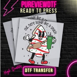 May include: DTF transfer featuring a cartoon Christmas tree character holding a green tumbler. The tree has red and white stripes and the text "out here lookin like a snack". The image includes the text "PUREVIEWDTF READY TO PRESS" and "DTF TRANSFER".