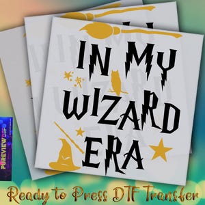 May include: White canvas with the text "IN MY WIZARD ERA" in black, with yellow accents including a broom, owl, stars, wand, and wizard hat. The phrase "Ready to Press DTF Transfer" is at the bottom.