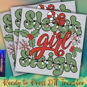 May include: Three stacked DTF transfer sheets with a Christmas design. The design features the words "Sleigh Girl Sleigh" in green and red, decorated with holly, flowers, and lights. The phrase "Ready to Press DTF Transfer" is at the bottom.
