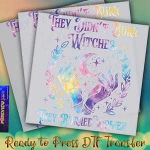 May include: Stacked white DTF transfer sheets with the text "THEY DIDN'T BURN WITCHES THEY BURIED WOMEN" in holographic foil. The design features hands, a crescent moon, and stars. The words "Ready to Press DTF Transfer" are at the bottom.
