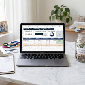 May include: A laptop displaying a nonprofit fundraising budget tracker sits on a white marble surface. A glass jar labeled "Community Fund - Thank You!" is filled with coins and bills. A clipboard, volunteer badge, and framed photo are also visible.