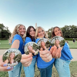 May include: Four friends wearing blue shirts with white text and images on them. The text on the shirts says "Avery's Big/Little". The friends are holding round buttons with images of people on them. The buttons also have text that says "Avery's Big/Little".