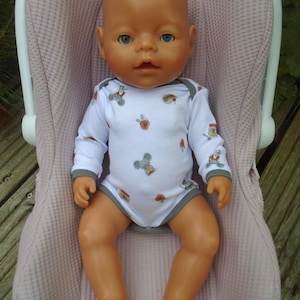May include: A bald baby doll with blue eyes and tan skin wearing a white long-sleeved bodysuit with a grey trim and a repeating pattern of small grey mice and orange jars. The doll is seated in a light pink textured infant car seat carrier.
