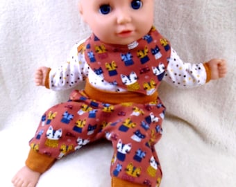 May include: A bald doll with blue eyes wearing a long-sleeved white shirt with small brown and black dots, a rust-coloured bib, and matching rust-coloured trousers. Both the bib and trousers feature a repeating pattern of stylised cats in blue, yellow, and white.