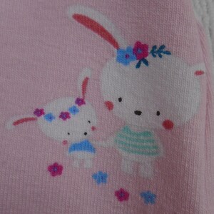 Puede incluir: Tela rosa con una ilustraci&oacute;n de dibujos animados de dos conejos blancos con coronas de flores. El conejo m&aacute;s grande lleva una camisa a rayas azules y verdes. Flores rosas, azules y moradas est&aacute;n esparcidas.