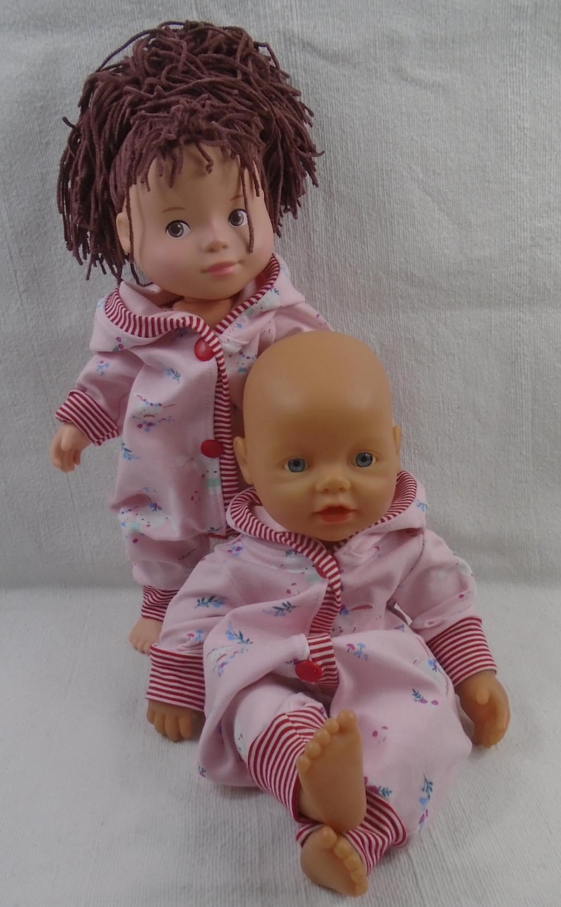 Puede incluir: Dos mu&ntilde;ecas beb&eacute;s con pijamas rosas a juego con estampado floral y ribetes a rayas rojas y blancas. Una mu&ntilde;eca tiene cabello casta&ntilde;o y la otra es calva. Las mu&ntilde;ecas est&aacute;n posadas sobre un fondo blanco.