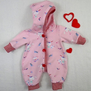Puede incluir: Mono rosa para beb&eacute; con capucha, con un estampado de conejitos blancos y detalles florales. El mono tiene ribetes a rayas rojas en los pu&ntilde;os, el forro de la capucha y la tapeta de los botones. Tres decoraciones rojas en forma de coraz&oacute;n est&aacute;n colocadas cerca del mono.