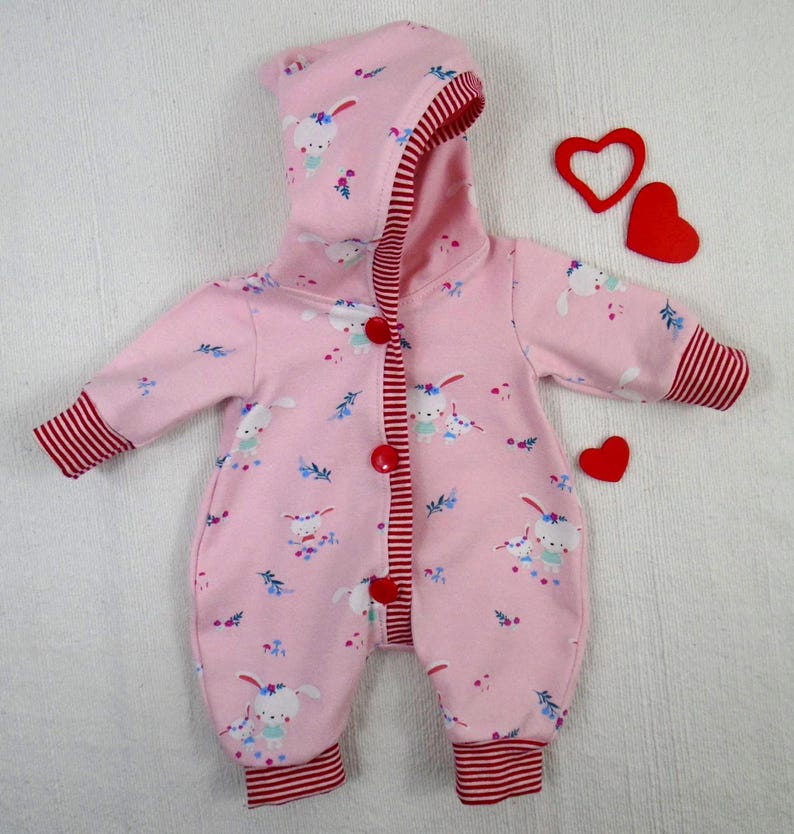 Puede incluir: Mono rosa para beb&eacute; con capucha, con un estampado de conejitos blancos y detalles florales. El mono tiene ribetes a rayas rojas en los pu&ntilde;os, el forro de la capucha y la tapeta de los botones. Tres decoraciones rojas en forma de coraz&oacute;n est&aacute;n colocadas cerca del mono.