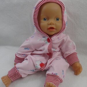 Puede incluir: Una mu&ntilde;eca beb&eacute; con un mono rosa con capucha y ribetes a rayas blancas y rojas. El mono presenta un estampado de conejitos blancos y detalles florales azules. La mu&ntilde;eca tiene ojos azules y est&aacute; sentada.