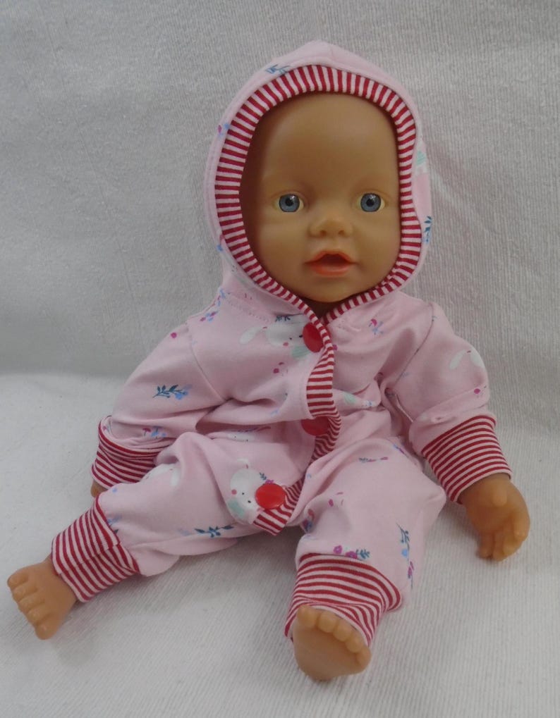 Puede incluir: Una mu&ntilde;eca beb&eacute; con un mono rosa con capucha y ribetes a rayas blancas y rojas. El mono presenta un estampado de conejitos blancos y detalles florales azules. La mu&ntilde;eca tiene ojos azules y est&aacute; sentada.