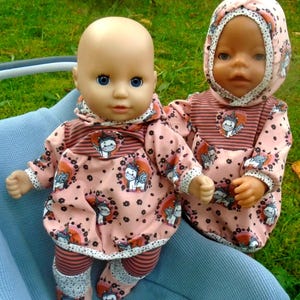 Könnte beinhalten: Zwei Babypuppen in passenden rosa Outfits mit Einhorn-Print. Die Outfits umfassen Kapuzenoberteile, gestreifte Leggings und Babyschuhe. Die Puppen haben blaue Augen und sitzen in einem blauen Kinderwagen.