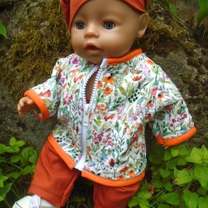 May include: A doll wearing a solid orange cap with a floral button, a white zip-up jacket featuring a colorful botanical print with orange trim, and solid orange pants. The doll has blue eyes and is wearing white sneakers with black stripes.