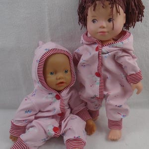 Puede incluir: Dos mu&ntilde;ecas con monos rosas con capucha a juego con ribetes a rayas rojas y blancas. Los monos tienen un peque&ntilde;o estampado floral y botones rojos. Una mu&ntilde;eca est&aacute; sentada, la otra de pie.